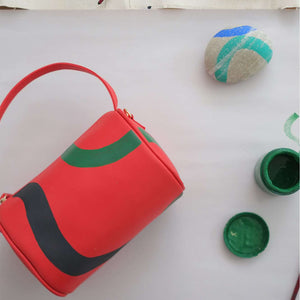 Vegan leather duffle style mini bag with detachable crossbody strap in red with hand painted squiggles in black and green.  Overhead shot wit art supplies.